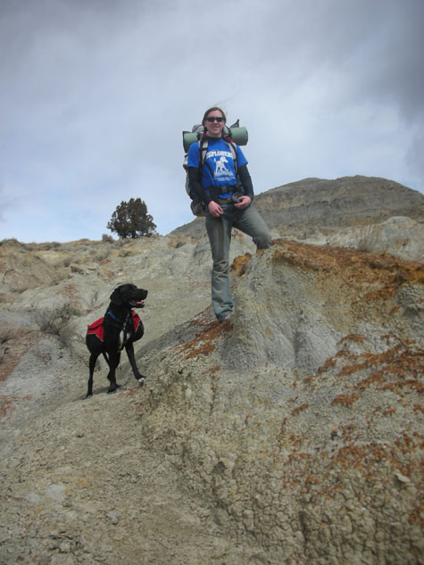 Ace the black lab mix wearing a backpack next to his owner while hiking