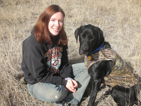 Woman sitting with black lab mix wearing hunting vest in a field